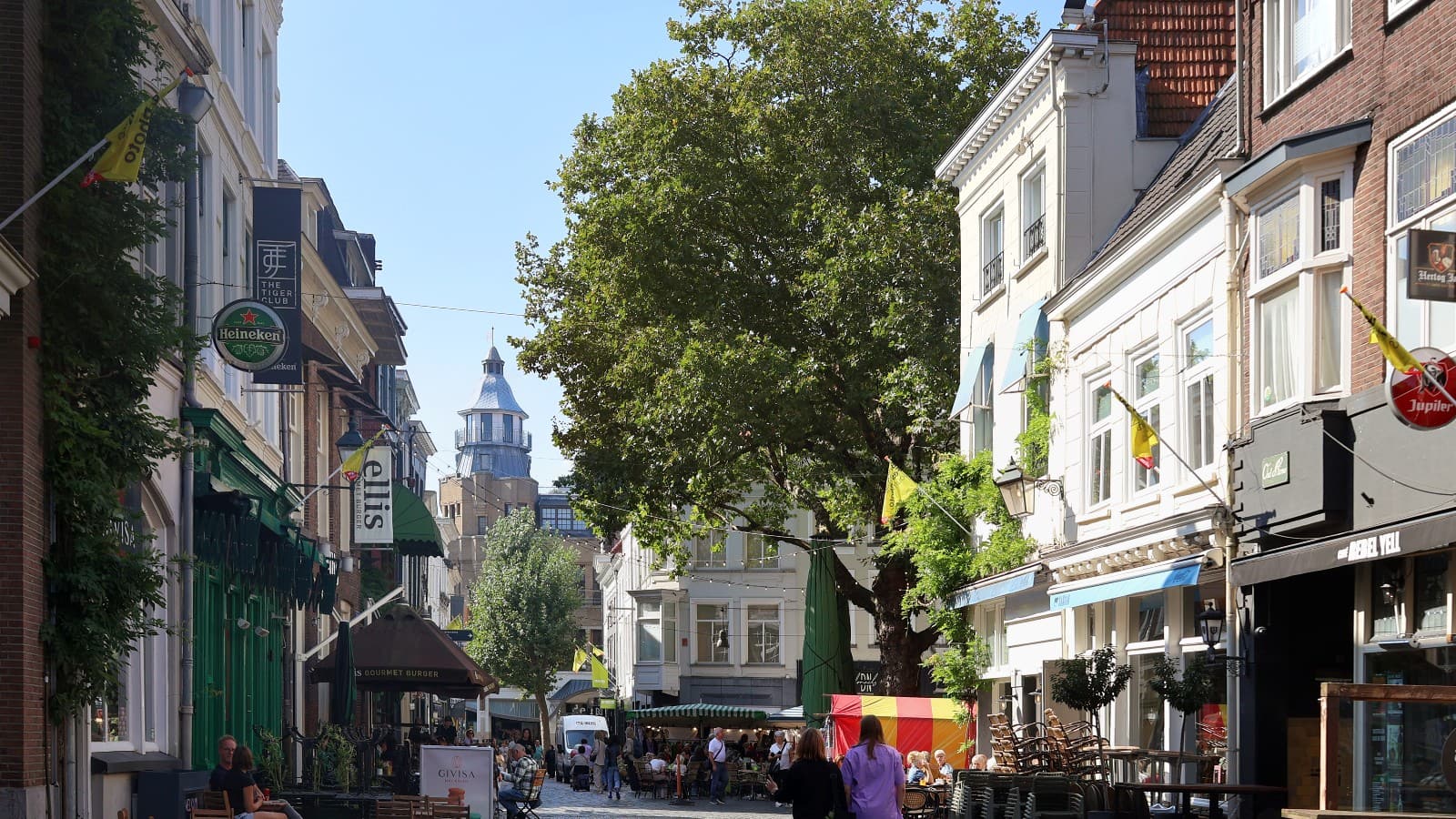 De Sint Janstraat in Breda met terrassen, winkels en een grote boom, met zicht op de Grote Markt.